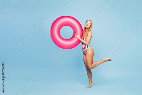 Full body sideways young sexy fun woman she wears bikini swimsuit rest near hotel pool hold in hand pink rubber ring look camera isolated on plain blue background. Summer vacation sea sun tan concept.