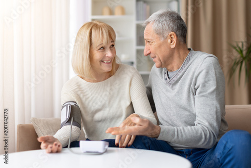 Smiling mature couple discussing health results after blood pressure check at home, happy elderly spouses showing care and positive wellness lifestyle indoors