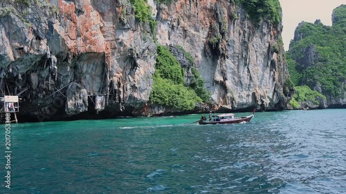 Traditional longtail boat with passengers navigating the clear turquoise waters, passing towering limestone karst cliffs covered in lush green tropical vegetation.