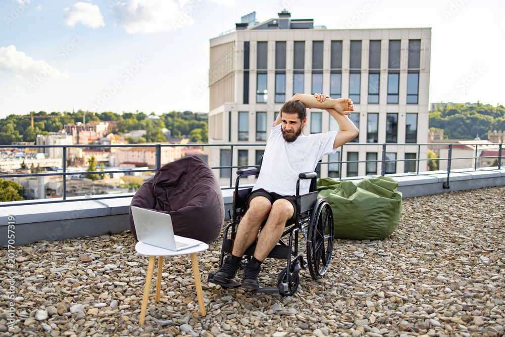 Fototapeta premium A man in a wheelchair stretches his arms on a rooftop terrace with a city view, beanbags, and a laptop nearby