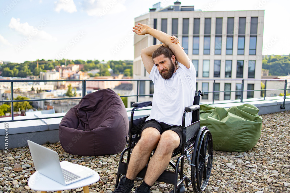 Fototapeta premium A man with a beard sits in a wheelchair on a rooftop terrace, stretching his arms while looking at a laptop on a small table