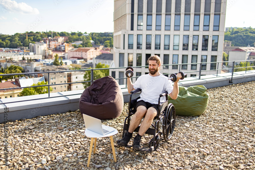 Obraz premium A man with a beard in a wheelchair is exercising with dumbbells on a rooftop terrace with a city view