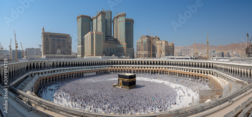 A high angle, panoramic shot captures a vast crowd of pilgrims dressed predominantly in white garments performing tawaf around the kaaba in the grand mosque.