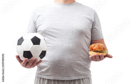 A fat male soccer fan in gray shorts and a t-shirt holds a soccer ball and a hamburger in his hand.