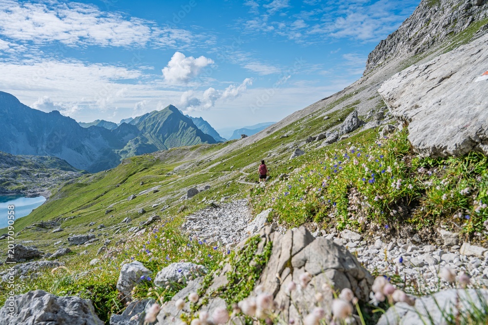 Obraz premium Female hiker with backpack and dog walking on mountain trail above Butzensee lake, Mohnenfluh and Braunarlspitze peaks in the background, Arlberg region, Vorarlberg, Austria