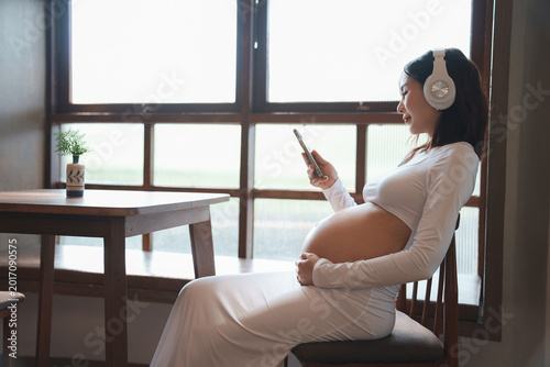 Pregnant woman sitting by window listening to music with headphones and mobile phone
