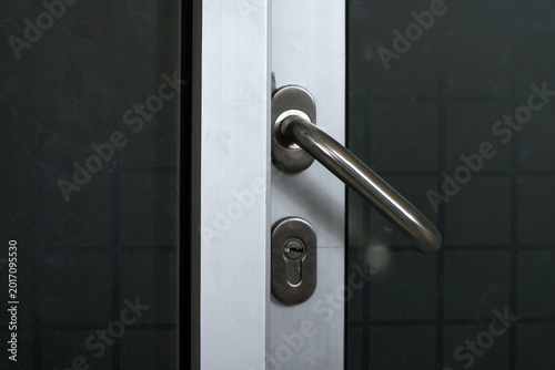 Close-up of a modern metallic door handle and keyhole lock on a silver aluminum frame. Minimalist glass door design for office or residential building, focusing on security and interior hardware detai