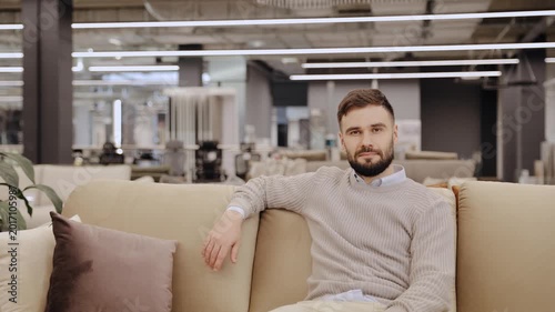 Young bearded man sitting relaxed on a beige sofa in a modern furniture showroom, looking at camera