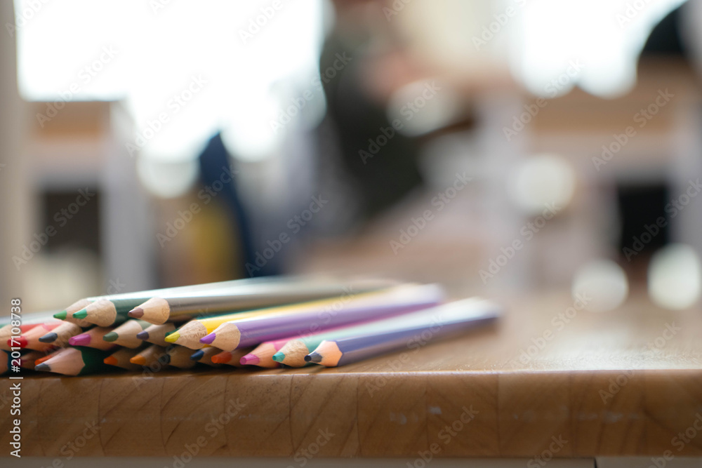 Fototapeta premium Colored pencils arranged on a wooden table with soft blurred classroom background