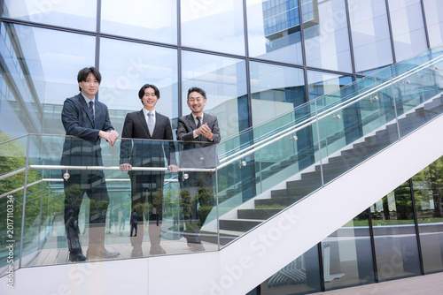 Bild auf Leinwand 屋外のビル背景で未来を見据え笑顔で語る会社のビジネスマンチームの成長と挑戦　プロジェクトの成功などのポジティブ前向き上司、若手の部下を見守るなど3人チームのイメ
