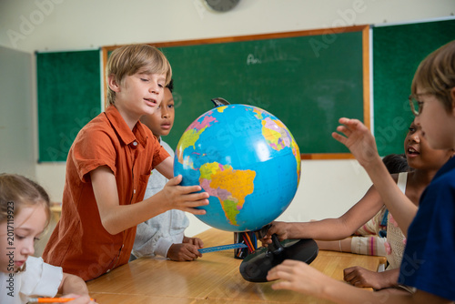 Primary schoolchildren looking at globe in classroom. Diverse children painting in classroom. Creativity and cultural exchange through art. International education and global knowledge sharing.
