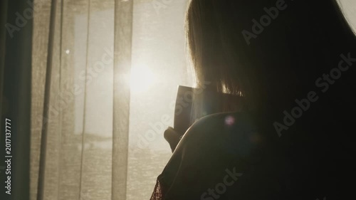 Silhouette of woman holding mug looking out window at sunrise.