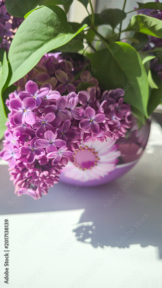Obraz premium A bouquet of blooming lilacs with green leaves in a round glass vase