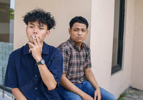 Young Asian Man Smoking Cigarette While Friend Looks At Him With Annoyed Expression