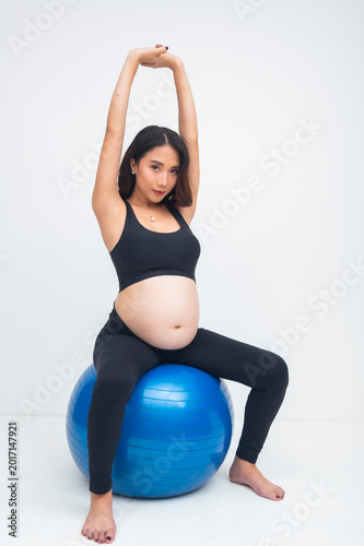 Quadro su tela Focused young pregnant Asian woman performing overhand shoulder stretch on a swiss ball, isolated on a white background during prenatal fitness workout