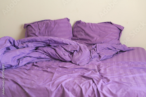 Fotografija A rumpled bed with purple sheets and pillows sits in a simple room