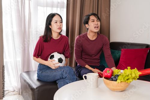 Tableau sur toile Asian couple watching a soccer match at home, sitting anxiously on a couch with