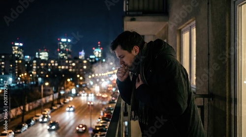 Sick young man in a heavy jacket coughing on an apartment balcony at night with city lights background.