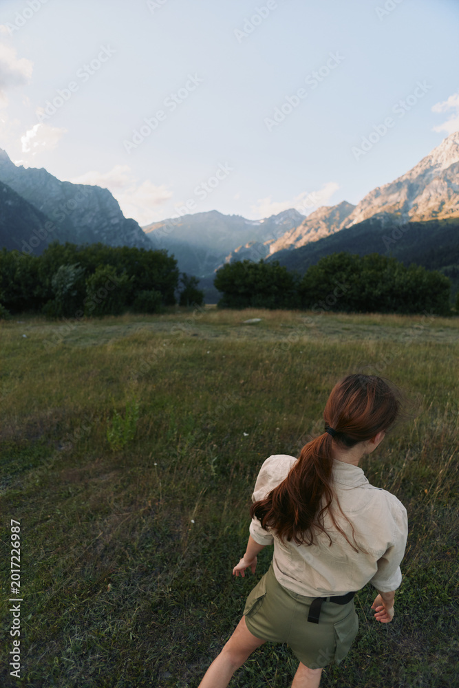 Obraz premium Young woman with long brown hair walks through a grassy field surrounded by mountains during a clear day, enjoying a peaceful outdoor lifestyle moment.