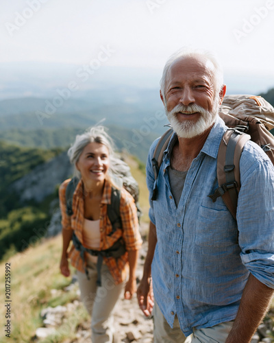 Sunlit trail winds through green hills. Senior couple hikes with smiles and backpacks. They move forward, enjoying the mountain air. Natural light highlights their joyful expressions