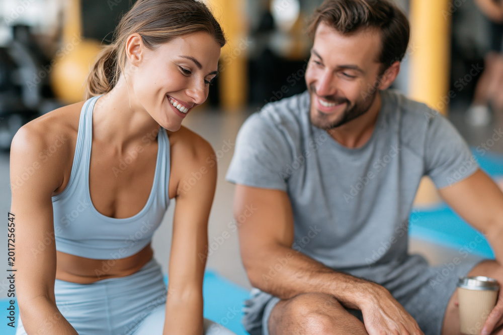 Fototapeta premium Young couple laughs together on yoga mats. They smile warmly, enjoying post-workout rest. Background hints at a modern fitness studio. Ideal for wellness, fitness, or healthy lifestyle content