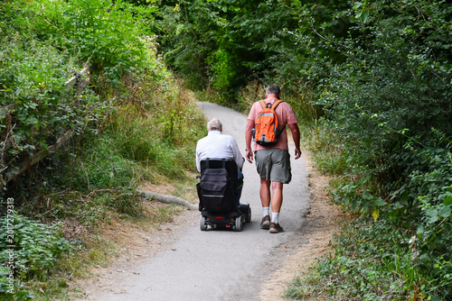 Fotografie Wheel chair walk, man on mobility scooter out enjoying the English countryside with his able bodied friend as they walk and chat together on a summers day