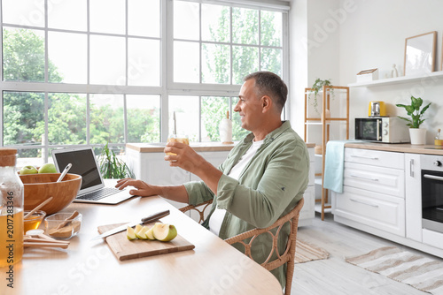 Mature man with apple cider using laptop at table in kitchen