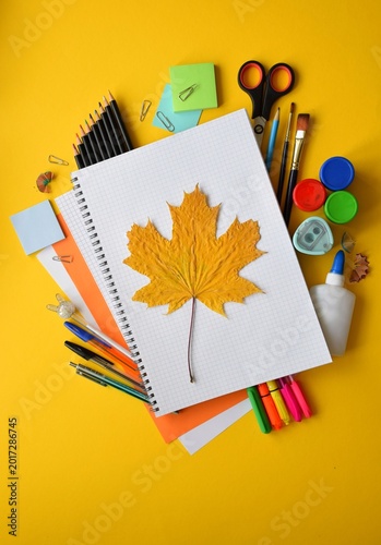 Autumn Maple Leaf on Open Notebook Surrounded by Colorful School Supplies on Yellow Background