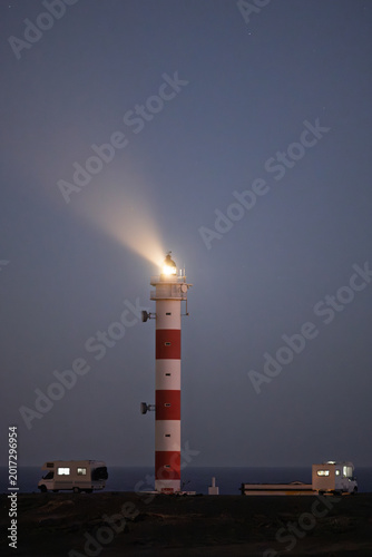 Red and white lighthouse glowing at night with camper vans
