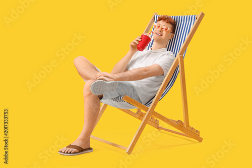 Young man with cup of soda resting in deck chair on yellow background