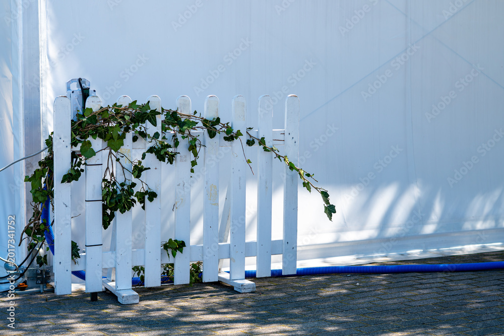 Ταπετσαρία White wooden picket fence entwined with ivy stands on a paved surface in front of a large white tent wall, with bright sunlight casting shadows and a blue technical hose