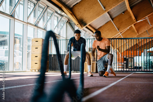 African American instructor offering encouragement to a fit young black man working out with battle ropes during an exercise session at the gym
