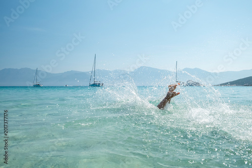 Young boy's arm splashing in clear turquoise sea of iconic Saleccia beach, with sailboats and mountains, evoking summer travel and relaxation at Saleccia and Lotu beach, Corsica.