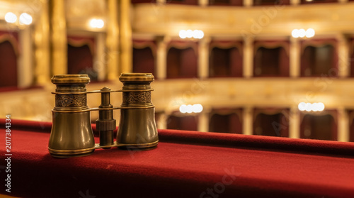 Conceptual macro shot of classic brass opera glasses resting on a plush deep-red velvet balcony railing in a grand theatre hall. Artistic patronage and high society concept.