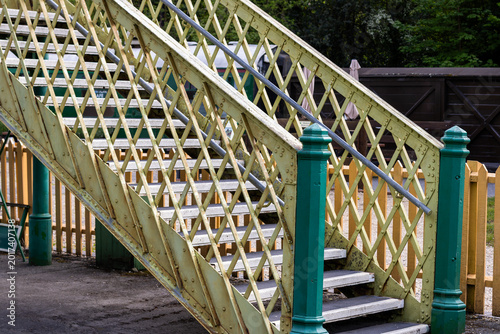 Foto Close-up view of a vintage pedestrian footbridge at a heritage railway station, featuring painted lattice metal railings, stairs, green posts, and classic station architecture