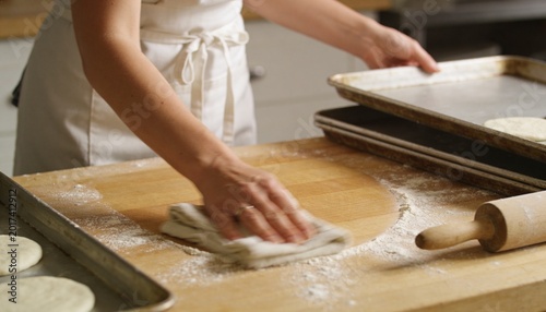 Wiping flour with cloth, baker in light apron spreading flour on workboard, rolling pin and sheets