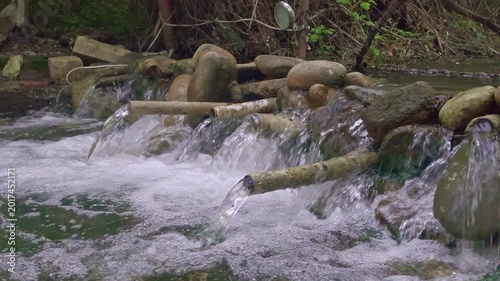 A serene close-up of a handmade stone water cascade in a lush green forest. Clear water flows through metal pipes into a bubbling stream, creating a tranquil and natural atmosphere. Ideal for wellnes