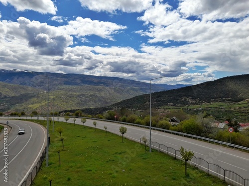 Panoramic High-Angle View of a Curving Mountain Highway near Koyulhisar, Turkey, with a Driving Car, Rolling Hills, Scattered Villages, and a Dramatic Cloudy Sky on a Bright Sprin