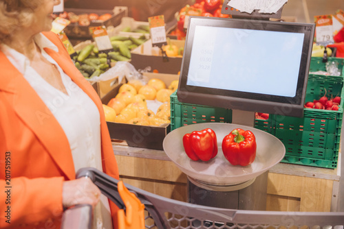 Slika na platnu Senior woman weighing red bell peppers on a scale, grocery shopping for fresh pr