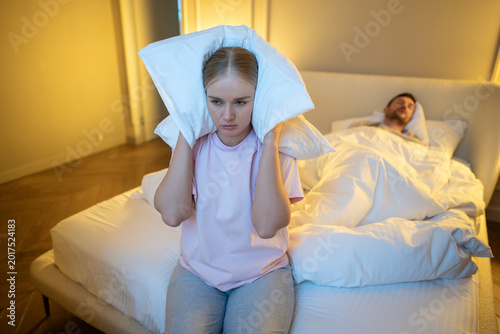 Photos Annoyed woman suffering from insomnia and frustration caused by husband snoring loudly at night, irritated wife sitting on bed covering ears with pillow to block snore sound while man sleeping