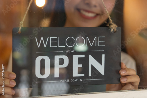 beautiful female business retail owner in medical face mask hanging open wooden sign board at the entrance door of the shop and ready to service customer.