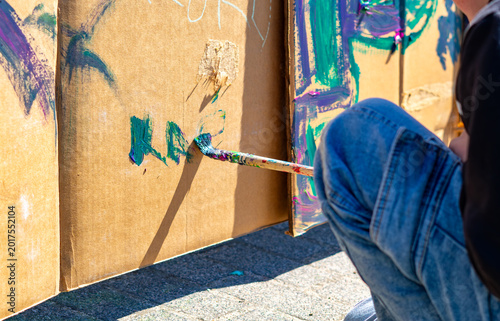 Close-up of a person painting with a brush on a large cardboard sheet outdoors, hand holding a brush with blue paint, person in jeans sitting on pavement, background with colorful paint splashes and g