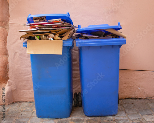 Two blue recycling bins overflowing with cardboard and paper waste against a neutral wall, representing waste management and environmental recycling efforts
