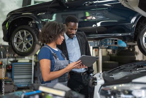 Female mechanic pointing at engine parts while holding a tablet displaying diagnostic data, explaining the repair to a male client.