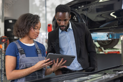 Female mechanic in a professional workshop uses a tablet to review vehicle maintenance checklist together with a client while inspecting the engine.
