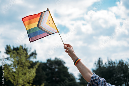 Hand raises progress pride flag against cloudy sky. Human rights advocacy, social justice movement, LGBTQ equality protest.
