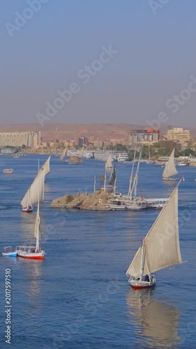 Aswan waterfront, Egypt, overlooking Nile River with traditional sailing feluccas, motor tourist boats, Elephantine Island and distant sand dunes under hazy sky.