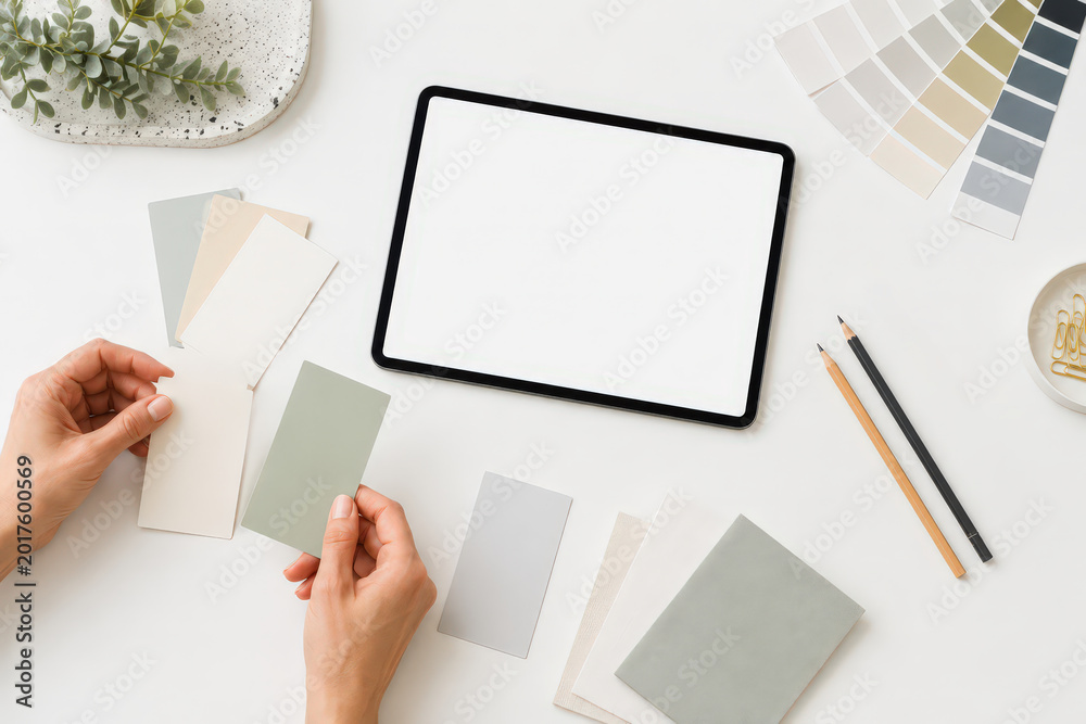 Obraz premium A person holds color samples while making choices for a home design project. A tablet sits on the table with empty space for notes. Pencils and additional color swatches are nearby.
