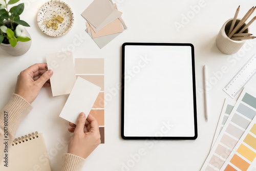 Two hands hold paint samples while a tablet sits on a table. A plant in a pot and a cup of pens are present. Color cards and a ruler are nearby. This setup shows a creative workspace.