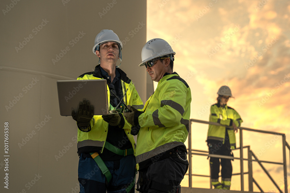 Fototapeta premium Male renewable energy engineers using AI algorithms on laptop to optimize wind turbine performance. Accelerating sustainable power generation at sunset.
